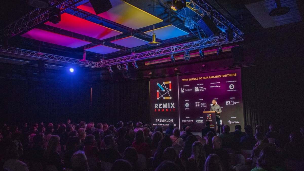 REMIX London. A gathering of arts, government, technology and media professionals sit watching a well dressed male speaker at a podium on a brightly lit stage. A large banner behind the speaker features the REMIX logo and the logos of the event's many sponsors and supporters.