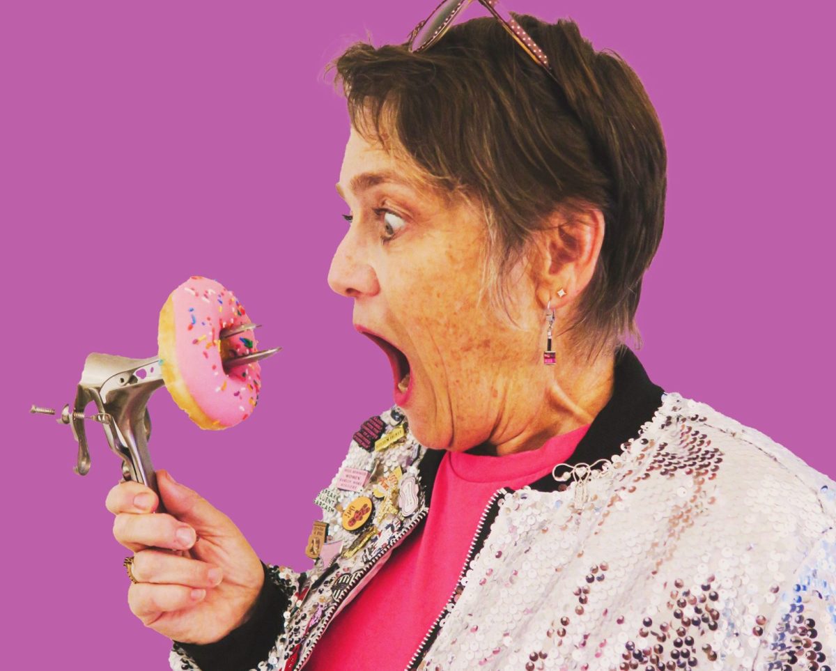 Against a pink backdrop a middle-aged woman with short hair, a bright pink T shirt and a glittery jacket is looking at a pink glazed donut which has the end of a speculum coming through it. The woman has her mouth wide open in feigned horror.