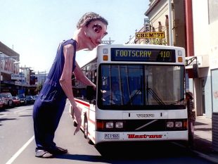 A giant male puppet looming over a bus that has Footscray on it.