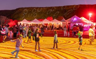 A group of kids standing on the red sand that has been projected with yellow and red circular patterns, while adults are watching on the side.