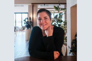 Vale Georgia Malone: a portrait photo of a female woman with white skin and short dark brown hair, sitting at a table with one hand under her chin. There are indoor plants in the background.