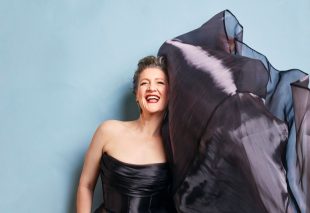 Virginia Gay, Adelaide Cabaret Festival 2025 Artistic Director. Gay, a fair-skinned woman, poses against a light blue backdrop and laughs as the elborate train of her black silk or satin gown billows up around her head, perhaps propelled by a wind machine out of the frame.