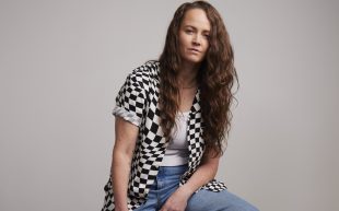 A Caucasian woman with long brown side parted hair wears a black and white checkered shirt over a white singlet and jeans. She is sitting on a stool. Zoë Coombs Marr.