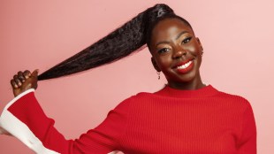 A black woman in a red top holding back her pony-tailed hair.
