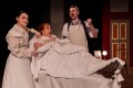 Dressed in Victorian era clothes, a woman in bed is surrounded by another women on her left and a man on her right. In the Next Room or The Vibrator Play