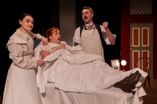 Dressed in Victorian era clothes, a woman in bed is surrounded by another women on her left and a man on her right. In the Next Room or The Vibrator Play