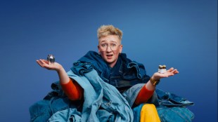 A blond bespectacled (trans) man surrounded by a pile of jeans.