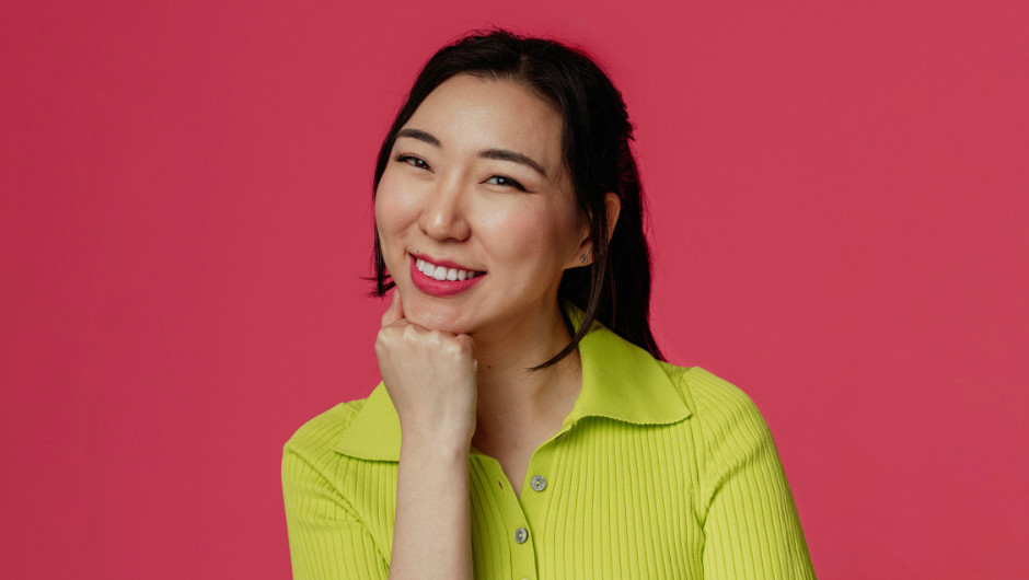 An Asian woman smiling against a pink background. She is wearing a yellow top.
