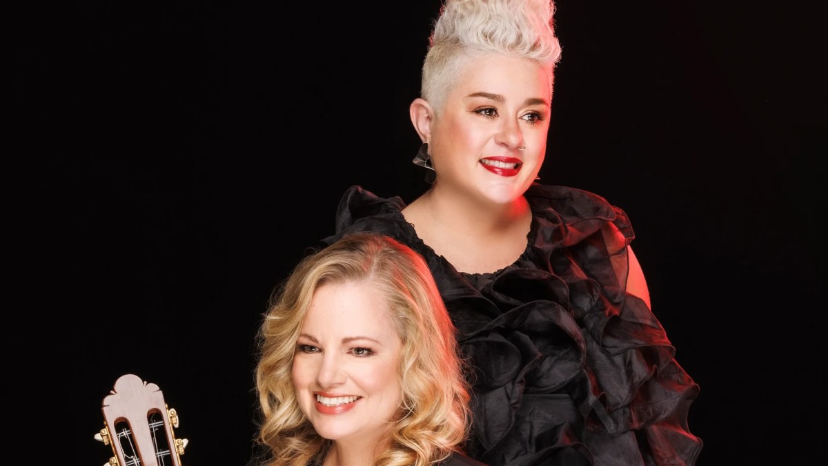 Two blonde women in black. The top of a guitar can be seen in the frame.