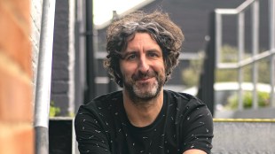 A man with wavy, brunette hair and a beard wearing a black top.
