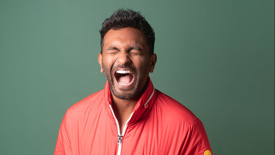 A Sri Lankan-Australian man with his mouth open and eyes closed. He's wearing an orangey red puffer jacket.