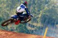 A motorbike is in the air riding through a forest and lifting off a dirt track.