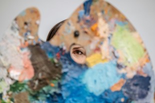 Picture of an artist's palette with paint splotches, and woman looking through hole in it. Arts news watch.