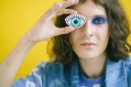 Woman with brown hair and heavy eye make up holding up drawn eye, yellow background. Arts news watch