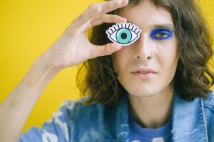 Woman with brown hair and heavy eye make up holding up drawn eye, yellow background. Arts news watch
