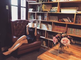 A woman is lying on a couch with a book over her face. There is a bookcase behind her and a coffee table on her right with flowers in a vase.