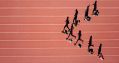 A group of people jogging on a running track; they are grouped in an arrow-head like formation, running towards the left of the photo. The image is taken from above, and shows their shadow stretched out beside them on the running track.
