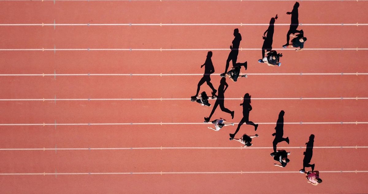 A group of people jogging on a running track; they are grouped in an arrow-head like formation, running towards the left of the photo. The image is taken from above, and shows their shadow stretched out beside them on the running track.