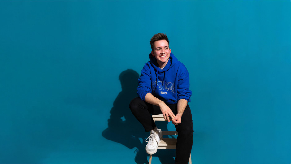 A (trans) man wearing a navy blue hoodie, white sneakers and black pants against a sky blue background.