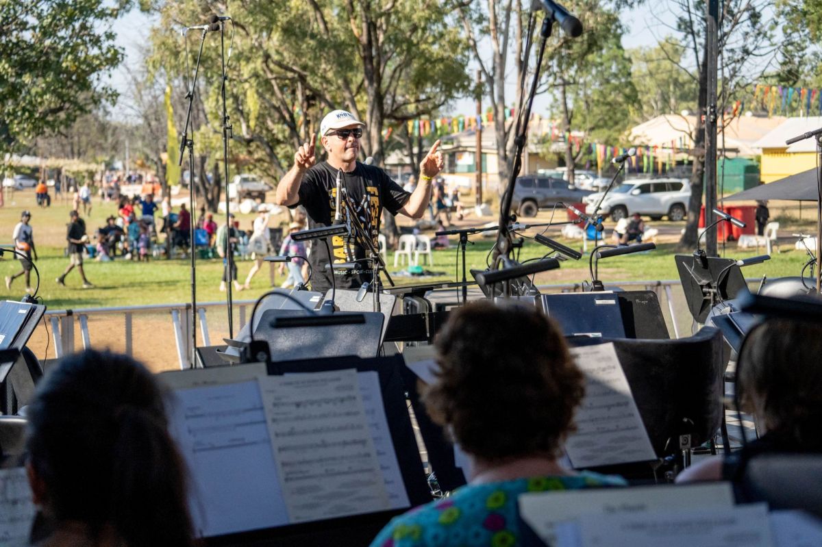 Darwin Symphony Orchestra (DSO) Territory Tour hero image. A casually dressed conductor, wearing a t-shirt and baseball cap, leads members of the DSO in an outdoor performance in a park. Cars, families on picnic rugs, and childfren playing footy on the grass are visible in the background.