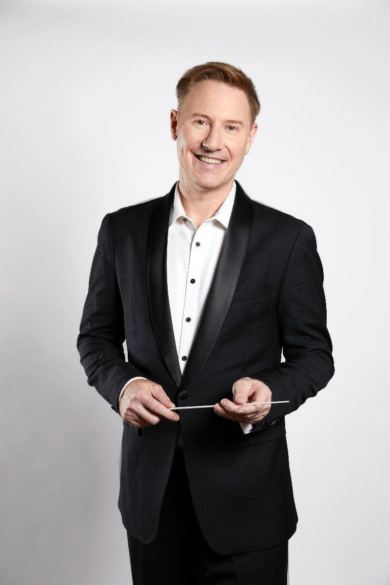 A caucasian man in a black suit with a white shirt and no tie holds a conductor's baton and smiles at the camera. MSO, Benjamin Northey.