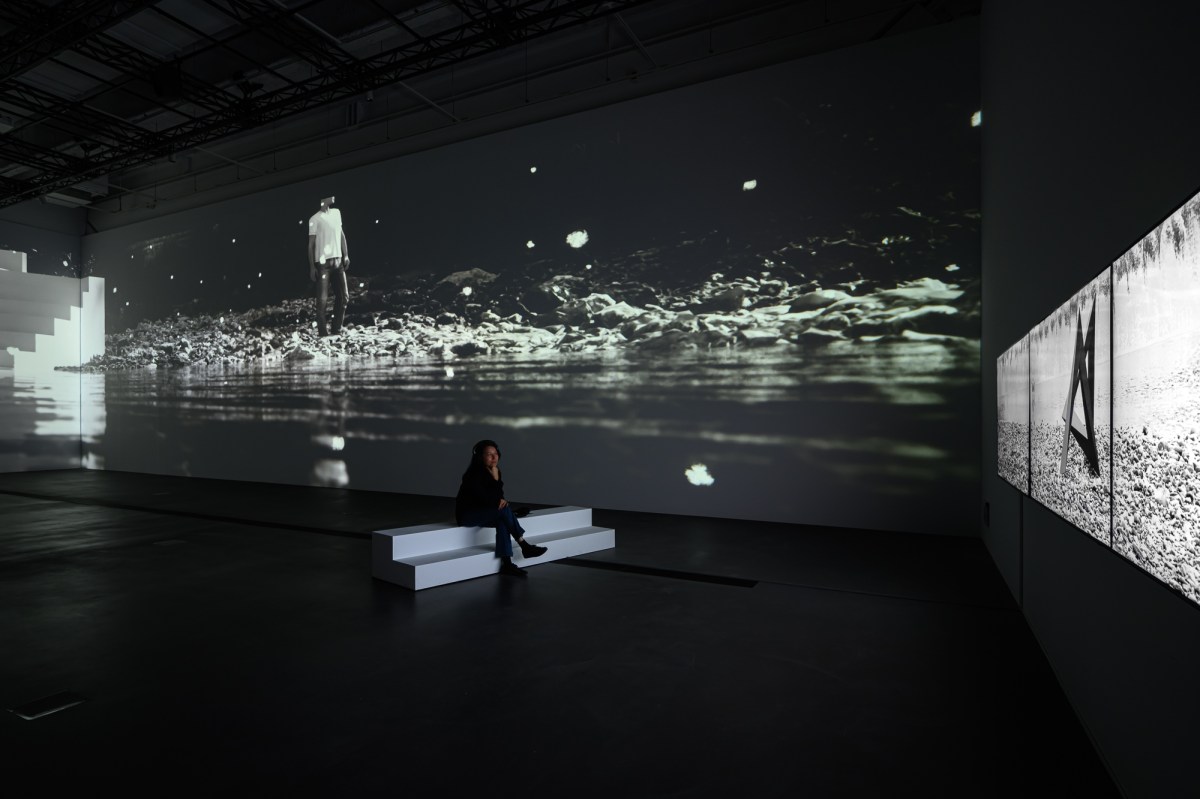 Large screens displaying black and white video inside a large dim gallery space. A single figure is sitting on a white step looking at one of the videos. 