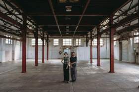 Vessel Contemporary: the interior view of a large empty industrial warehouse space with a young adult female and male person pictured standing together at the centre of the space.