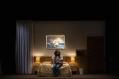 A Black woman is sitting on a bed spread which features a yellow and white design. The light is subdued.