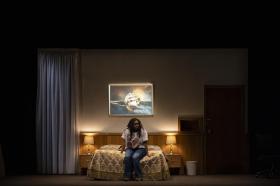 A Black woman is sitting on a bed spread which features a yellow and white design. The light is subdued.