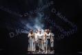 Six people dressed in varying costumes of white are huddle together on stage. In the background the words 'De-Kolo-nize' are spelt out diagonally.