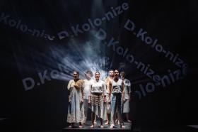 Six people dressed in varying costumes of white are huddle together on stage. In the background the words 'De-Kolo-nize' are spelt out diagonally.