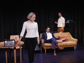 A blonde woman in a white top and black pants is standing near a clock that says 1978. A man is sitting on a brown couch and behind him is another man standing in the distance. A scene from Thursday’s Child Theatre's 2025 production of Harold Pinter's 'Betrayal'.