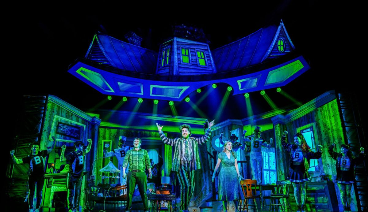 A scene from the Australian production of 'Beetlejuice The Musical'. A cartoonish haunted house primarily lit in purple and green; Eddie Perfect as 'bio-exorcist' Beetlejuice, stand with hands raised centre stage. He has green hair and a green and purple-striped suit. Other charactors, including a startled looking couple and members of the ensemble dressed as male and female cheerleaders stand onstage with them and behind them.