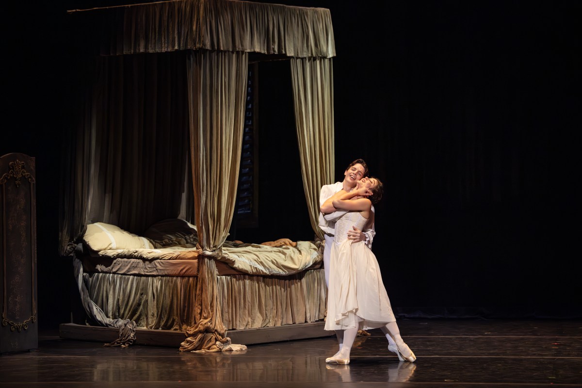 A couple in white embrace beside a bed with heavy drapes.