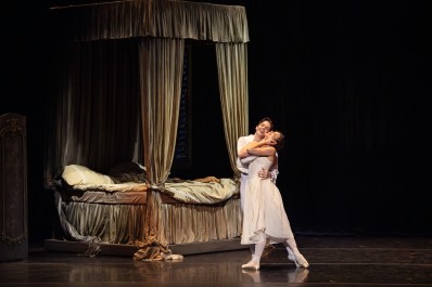 A couple in white embrace beside a bed with heavy drapes.