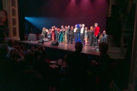 Approximately 15 artists take a bow on stage with audience members seated in the seats in front of them and also on the stage behind them. The photo depicts the final curtain at Perth International Cabaret Festival's closing gala in 2024.