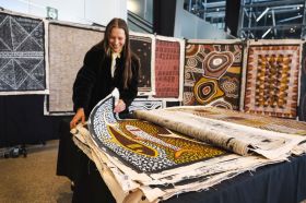 Artists from across the country will gather in Sydney for the National Indigenous Art Fair in July. The photo shows a woman looking through a stack of unframed Indigenous paintings on linen; more Indigenous artworks hang behind her.