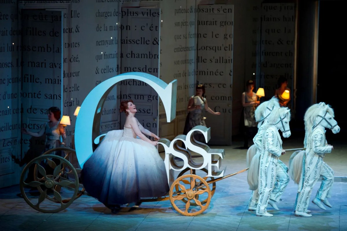 A production on stage with a woman sitting inside a carriage shaped like the letter ‘C’. Opera Australian production.
