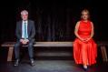 A man in a grey suit and a woman in a red dress are sitting apart on a park bench.