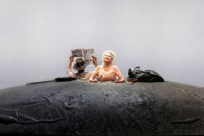 A scene from STC's 2025 production of Beckett's 'Happy Days'. A woman with her bottom half hidden in a mound of dirt. There's a black plastic bag next to her, and on her other side is man with his back to us, holding up a newspaper. He too is visible only from his waist up.