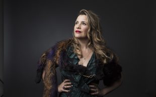 A profile photo of cabaret artist Ali McGregor, wearing a dark green dress with fur shoulder accents and flowy waves in her hair.