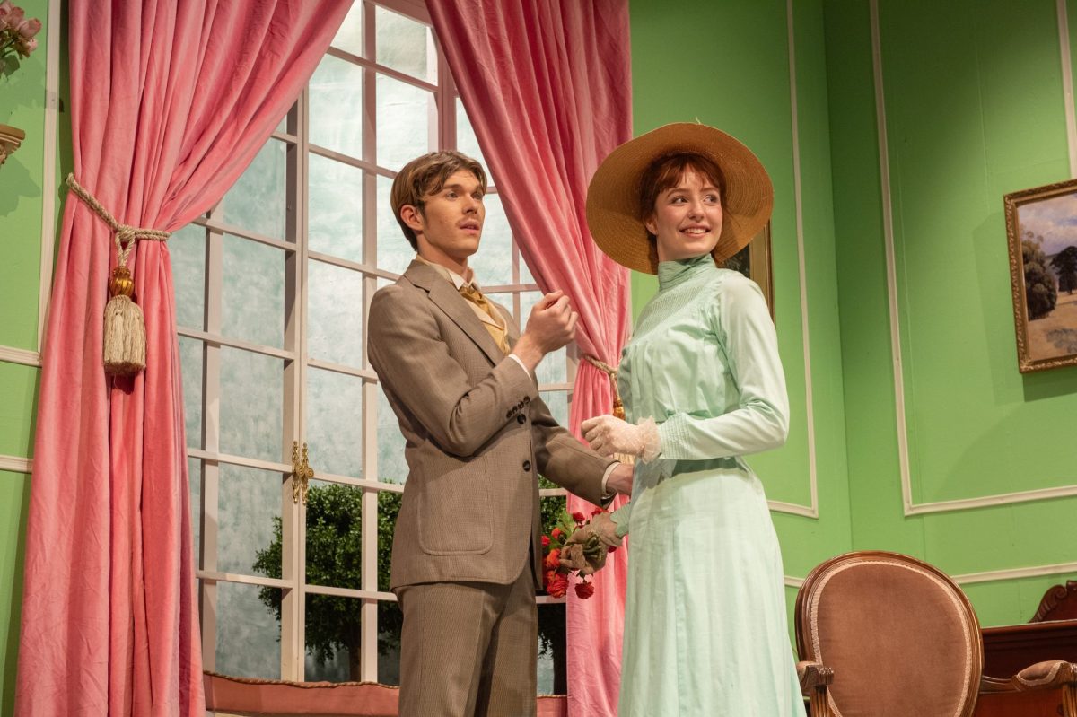 On a green and pink Edwardian stage set a young man in a brown suit and a young woman. in a pale green dress and large brimmed hat look off to the right. Lord Arthur Savile's Crime.
