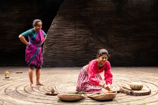 Two Indian women in colourful saris. One is standing, one is kneeling over some woven baskets.