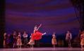 A ballerina in red photographed mid-leap. There are various cast members behind her.