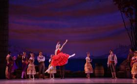 A ballerina in red photographed mid-leap. There are various cast members behind her.