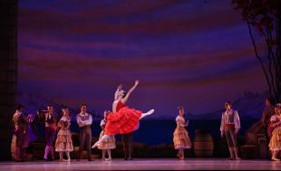 A ballerina in red photographed mid-leap. There are various cast members behind her.