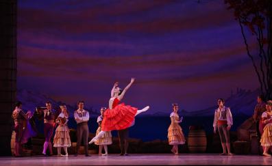 A ballerina in red photographed mid-leap. There are various cast members behind her.