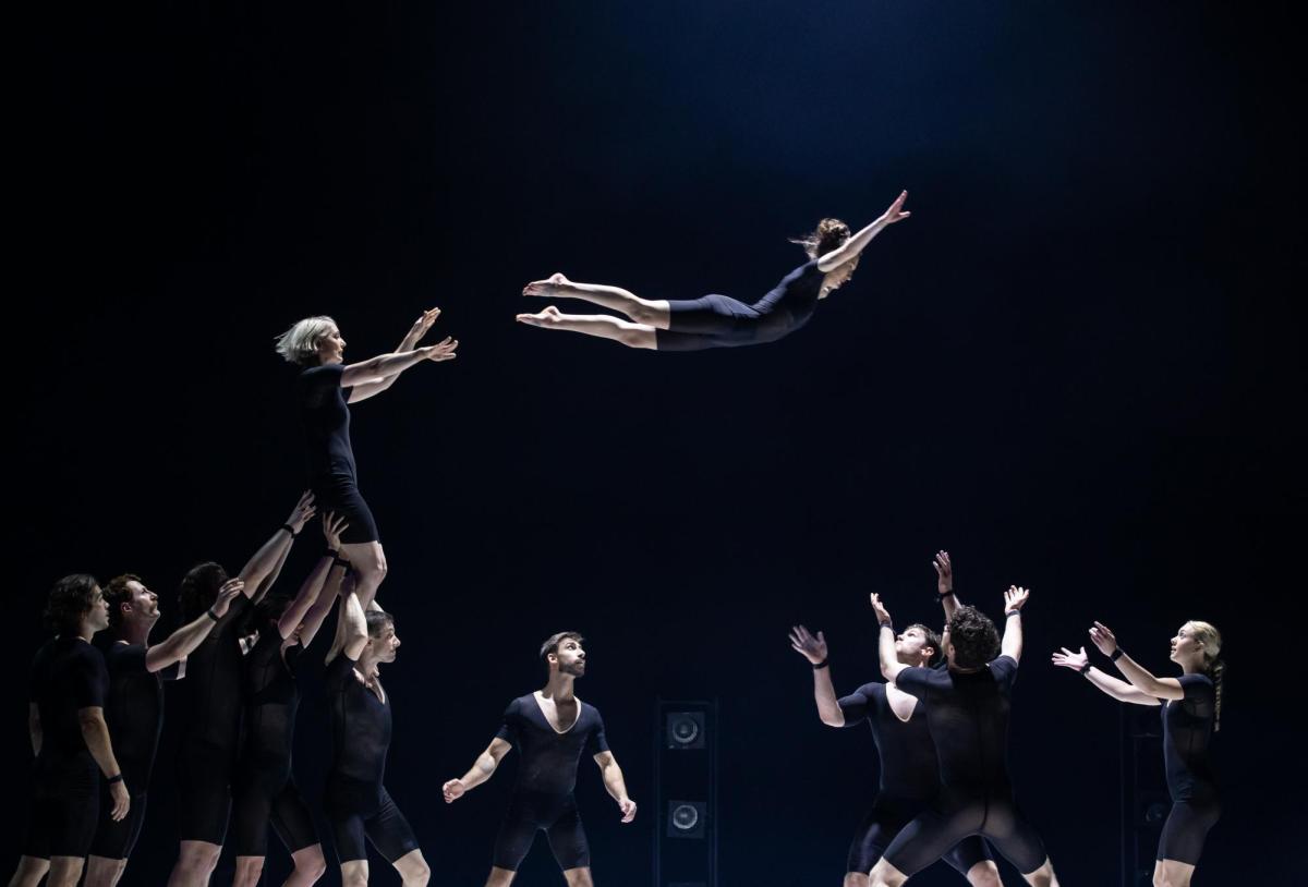 A group of circus performers in black leotards. One of them has been flung into the air and is flying towards more of the ensemble, who stand with arms raise to catch her. The photograph depicts a scene from Circa's 'Humans 2.0' at Arts Centre Melbourne.