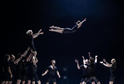 A group of circus performers in black leotards. One of them has been flung into the air and is flying towards more of the ensemble, who stand with arms raise to catch her. The photograph depicts a scene from Circa's 'Humans 2.0' at Arts Centre Melbourne.