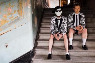 Two men sitting on a flight of white stairs. They are wearing shorts and white suit jackets adorned with palm trees. One of pair has a blank white mask, hat and sunglasses; the man to his left was a tanned face and a moustache.